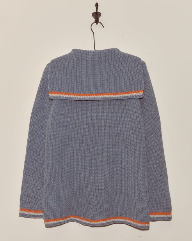 The Boat Deck Pullover, a blue virgin wool sweater with orange and white stripes, pirate ship buttons, and a sailor collar, hangs on a wall hook as seen from the back.