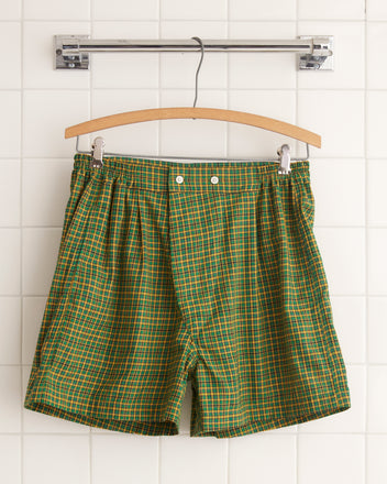 The Bobby Plaid Boxer Shorts in green and orange, made from midcentury shirting fabric, hang elegantly on a wooden hanger against a white tiled wall.