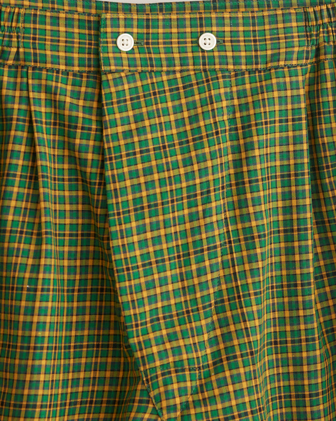 The Bobby Plaid Boxer Shorts in green and orange, made from midcentury shirting fabric, hang elegantly on a wooden hanger against a white tiled wall.