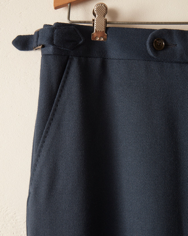 The Twill Side Buckle Trousers - Petrol hang on a wooden hanger against a light background, highlighting their belt and button details that evoke midcentury suiting style.