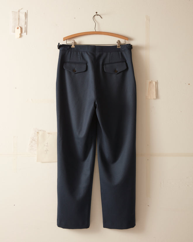 The Twill Side Buckle Trousers in Petrol, made from 100% wool, hang elegantly on a wooden hanger against a light wall.