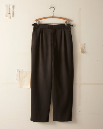 A pair of Twill Double Button Trousers - Black, made from 100% wool, hangs on a wooden hanger against a beige wall. Small pieces of paper are attached to the trousers, evoking the style of men's high-rise fashion from the 1930s-1940s.