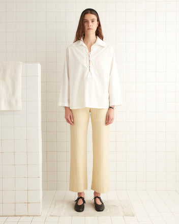 A person stands in a tiled room wearing a loose, 1960s-inspired Bonnie Shirt in Ecru and beige striped pants, with black shoes. The background includes a white towel on a partition wall.