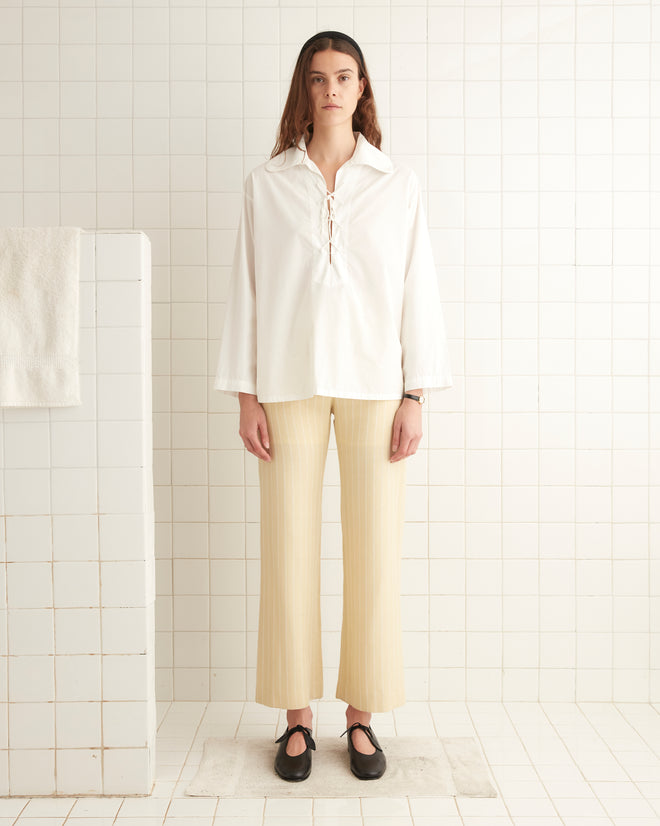 A person stands in a tiled room wearing a loose, 1960s-inspired Bonnie Shirt in Ecru and beige striped pants, with black shoes. The background includes a white towel on a partition wall.