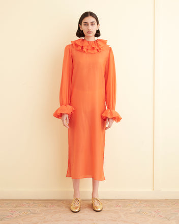 A person stands indoors against a cream-colored wall, wearing the Boo Ruffle Gown in orange, which features long sleeves and a ruffled collar. They pair the gown with gold shoes.