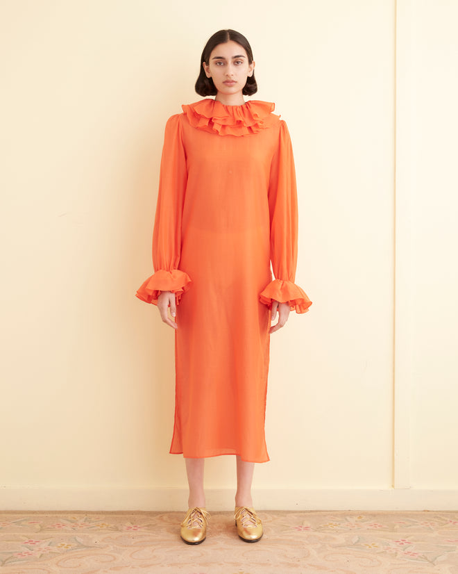 A person stands indoors against a cream-colored wall, wearing the Boo Ruffle Gown in orange, which features long sleeves and a ruffled collar. They pair the gown with gold shoes.