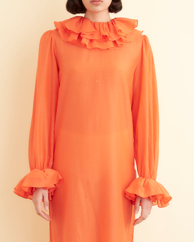 A person stands indoors against a cream-colored wall, wearing the Boo Ruffle Gown in orange, which features long sleeves and a ruffled collar. They pair the gown with gold shoes.