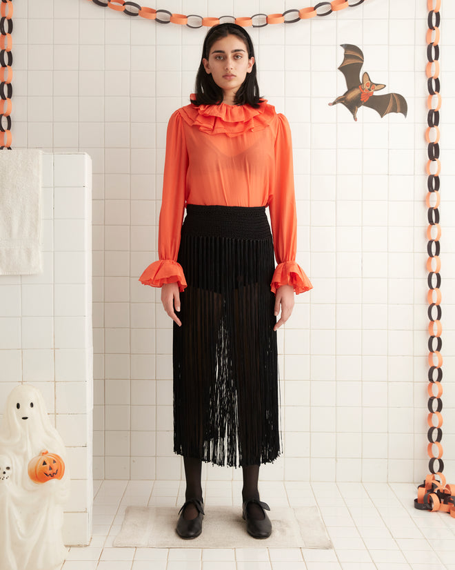 A woman stands against a pale yellow wall, wearing a 1970s inspired Boo Ruffle Blouse in orange, paired with white pants and black shoes.