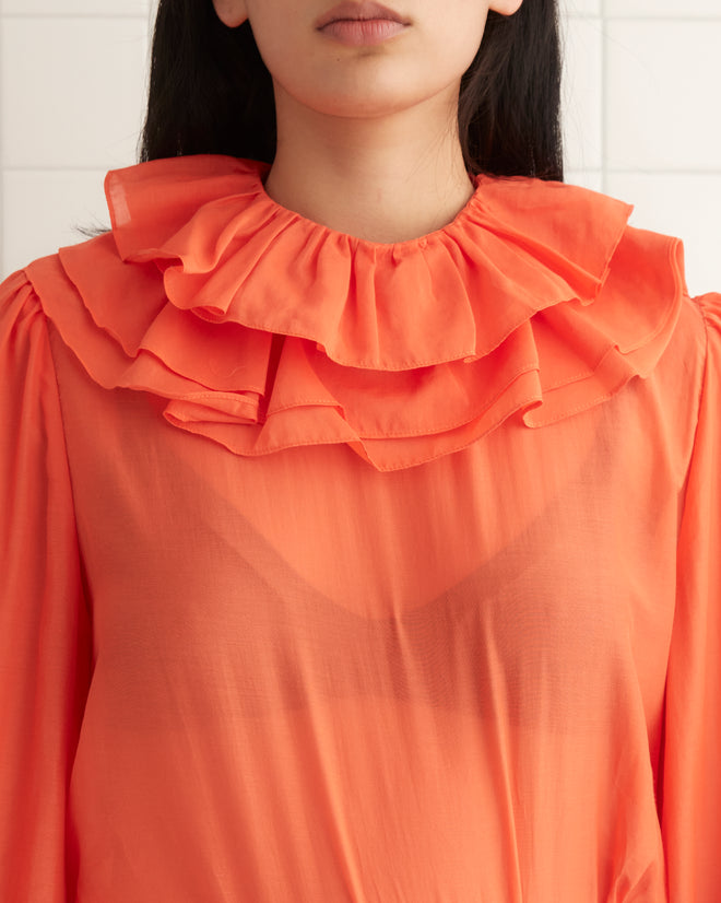 A woman stands against a pale yellow wall, wearing a 1970s inspired Boo Ruffle Blouse in orange, paired with white pants and black shoes.