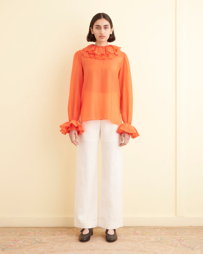 A woman stands against a pale yellow wall, wearing a 1970s inspired Boo Ruffle Blouse in orange, paired with white pants and black shoes.