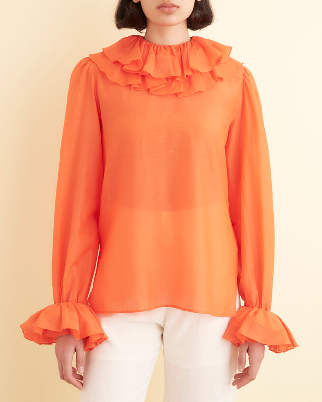 A woman stands against a pale yellow wall, wearing a 1970s inspired Boo Ruffle Blouse in orange, paired with white pants and black shoes.