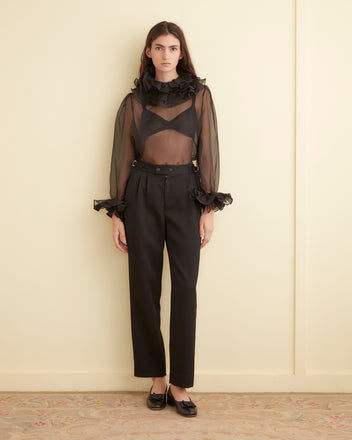 A person is elegantly dressed in the Boo Ruffle Top in sheer black and black trousers, standing gracefully in a minimalist room.