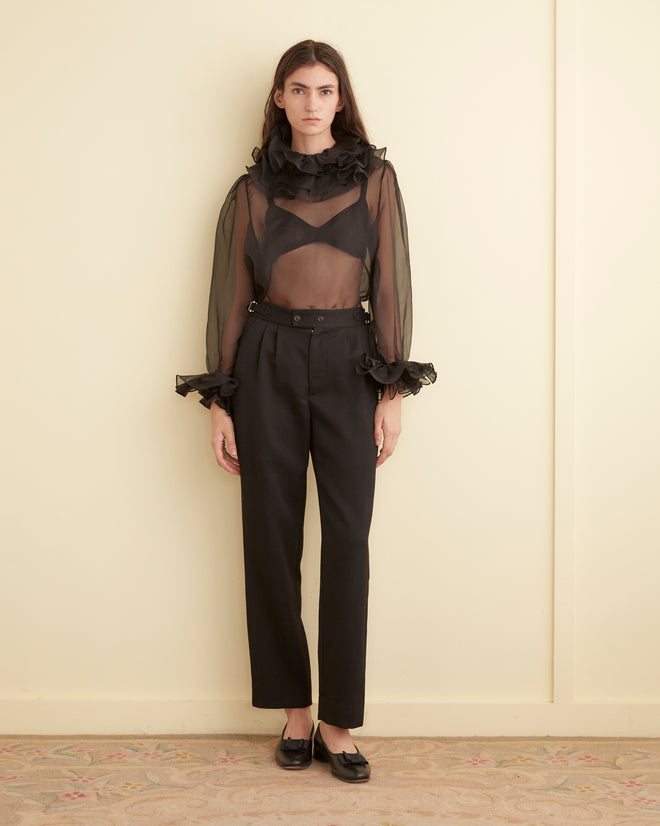 A person is elegantly dressed in the Boo Ruffle Top in sheer black and black trousers, standing gracefully in a minimalist room.