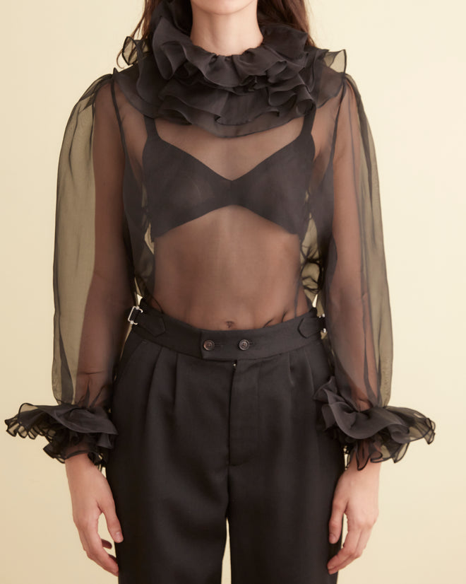 A person is elegantly dressed in the Boo Ruffle Top in sheer black and black trousers, standing gracefully in a minimalist room.