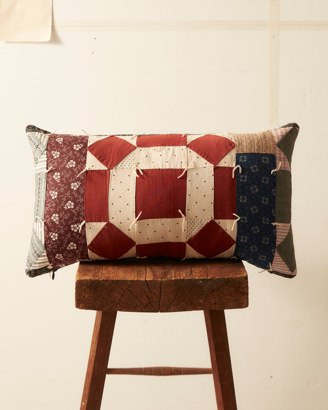 The Botanic Belvedere Pillow, crafted from a beautiful mix of calico prints and red, white, and blue patterned fabric, sits gracefully on a wooden stool in front of a plain wall.