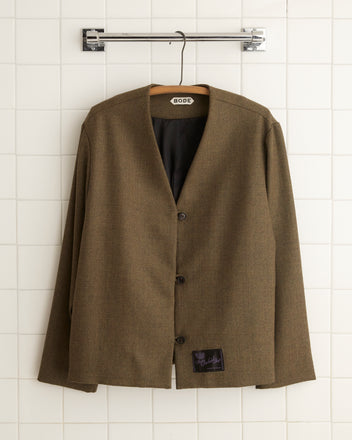 The Bottlebrush Jacket - 10 in brown wool, featuring a boxy fit, hangs elegantly on a hanger against a tiled wall backdrop.