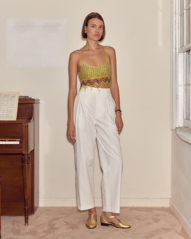Woman in a Bottleglass Top and white pants stands indoors by a piano and a window, looking at the camera.