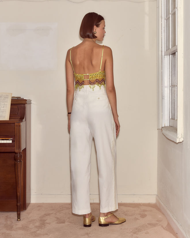 A woman stands indoors, facing away, wearing white pants, gold shoes, and the Bottleglass Top.