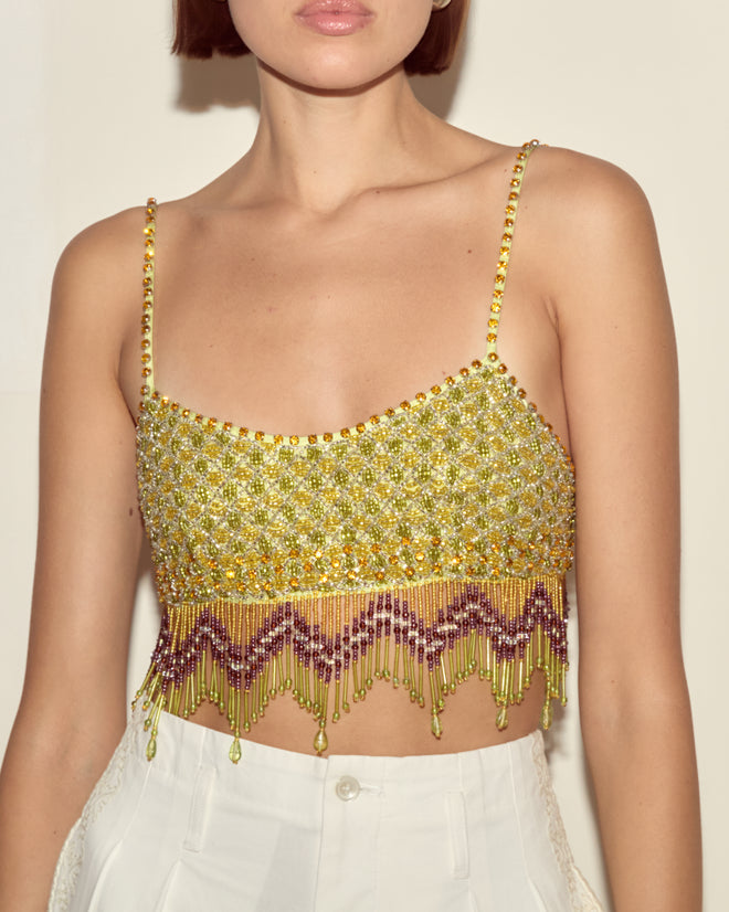 Woman wears the Bottleglass Top, a hand-beaded yellow crop top with fringe detail, paired with white high-waisted pants; torso view.