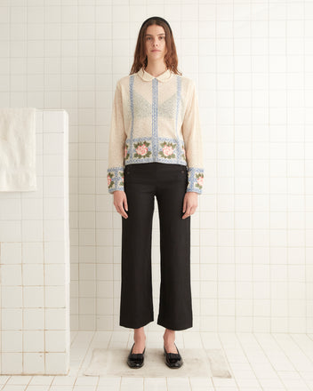 In a tiled room, a person stands wearing the 1920s inspired Bourbon Rose Blouse with floral patterns and black pants, with a folded towel visible in the background.