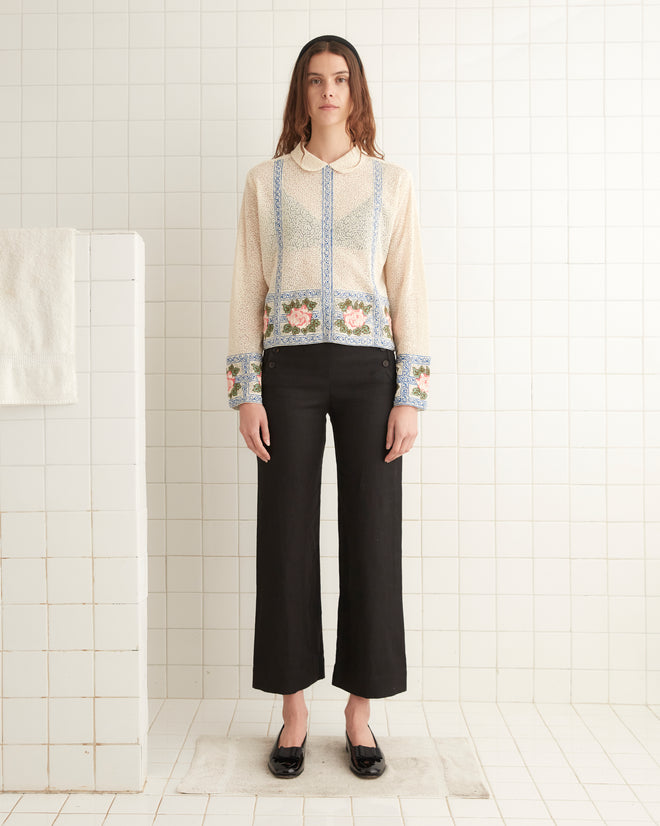 In a tiled room, a person stands wearing the 1920s inspired Bourbon Rose Blouse with floral patterns and black pants, with a folded towel visible in the background.