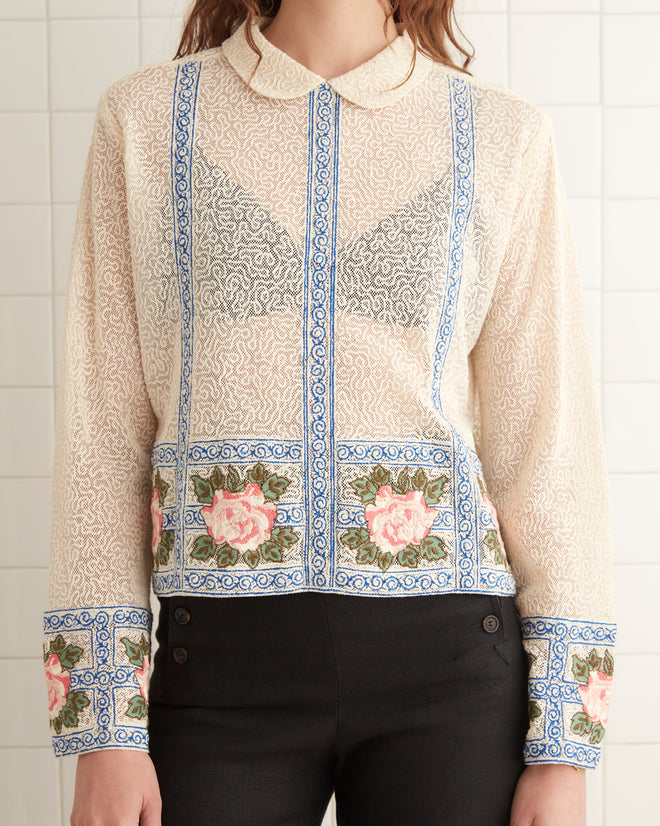 In a tiled room, a person stands wearing the 1920s inspired Bourbon Rose Blouse with floral patterns and black pants, with a folded towel visible in the background.