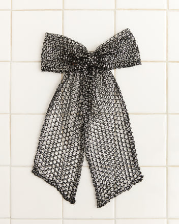 The Tanglenet Bow Barrette, a handmade black lace bow decorated with silver sequins, is elegantly attached to a tiled white wall.