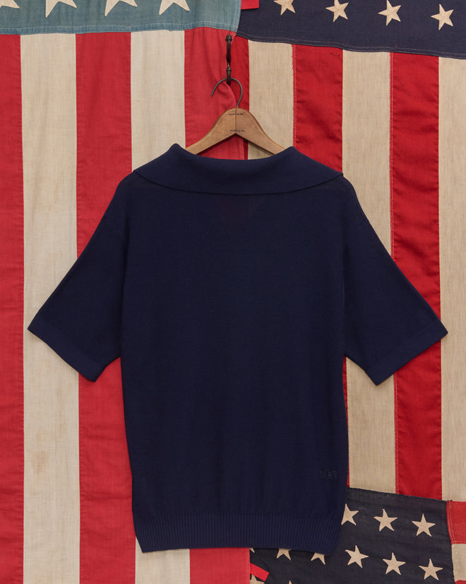 The Boxy Polo - Navy, a cotton knit short-sleeve sweater, hangs on a wooden hanger with an American flag in the background.