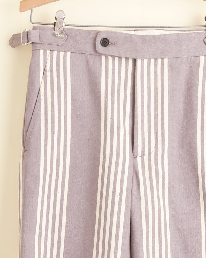 A pair of Boysenberry Stripe Trousers, featuring a light gray and white vertical stripe pattern reminiscent of deadstock ticking trousers, hanging on a wooden hanger against a light-colored wall. The side-buckle waist adjusters add a functional touch to these timeless classics.