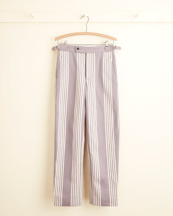 A pair of Boysenberry Stripe Trousers, featuring a light gray and white vertical stripe pattern reminiscent of deadstock ticking trousers, hanging on a wooden hanger against a light-colored wall. The side-buckle waist adjusters add a functional touch to these timeless classics.