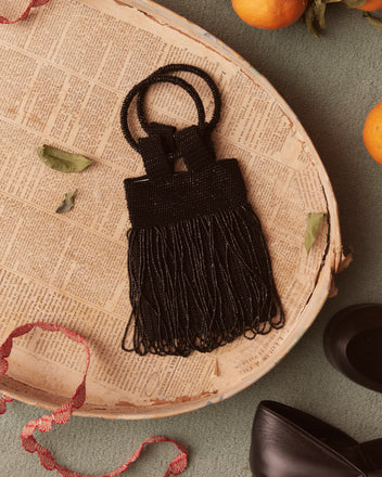 The Bracelet Evening Bag - Black, a beaded evening purse with a ring handle, rests on a vintage tray surrounded by scattered leaves, a shoe, ribbon, and oranges. This stunning piece captures the timeless elegance of a 1930s reproduction.