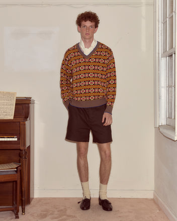 A man in a Bracken Sweater, collared shirt, dark shorts, and loafers stands by a piano in a sunlit room.