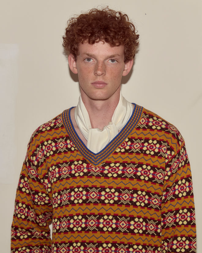 A young man with curly red hair wears the Bracken Sweater and a white scarf, standing against a plain wall.