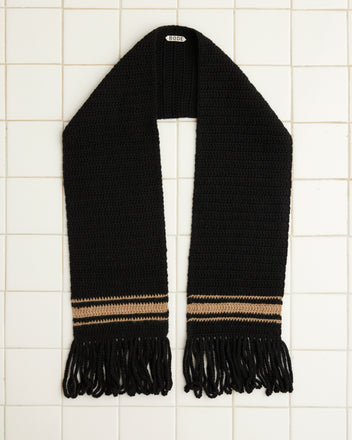 The Braided Fringe Scarf, hand-crocheted from wool in black with brown and tan stripes and finished with fringe ends, lies elegantly on a tiled surface.