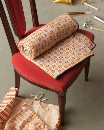 The Floral Jacquard Throw in Marigold rests elegantly on a red cushioned chair, accompanied by a beige bag and ribbon on the floor, perfect for enhancing your home goods collection with its cozy patterned design.