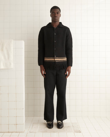 A man stands in a tiled room, wearing the Braided Fringe Shirt, paired with black pants and black shoes. A towel hangs on a low partition to his left. His elongated fit showcases an elegant silhouette that's both timeless and modern.