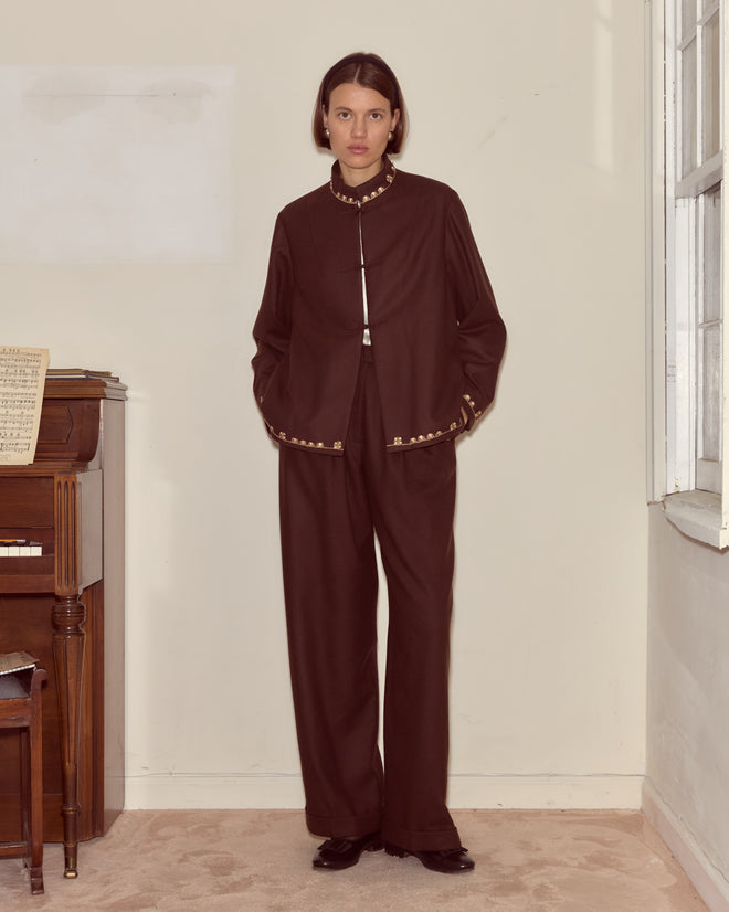 A woman stands indoors beside a piano, looking directly at the camera, wearing the Bramble Jacket in dark brown wool twill and classic suiting.
