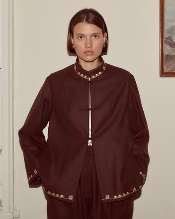 A woman stands indoors facing the camera with her hands in the pockets of a brown Bramble Jacket featuring gold trim.