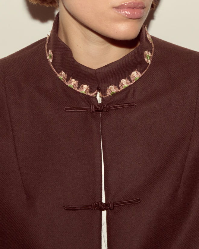 A person wearing the Bramble Jacket, a maroon swing style with frog closures, classic suiting details, and floral embroidery on the collar.