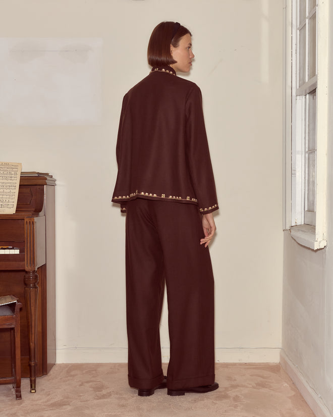 A woman wearing the Bramble Jacket in dark brown wool twill stands near a piano, facing away in a softly lit room.