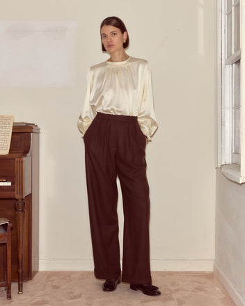 A woman stands indoors by a piano and window in classic style, wearing a cream silk blouse paired with Bramble Port Trousers—high-waisted, wide-leg, and dark—embodying timeless suiting elegance.