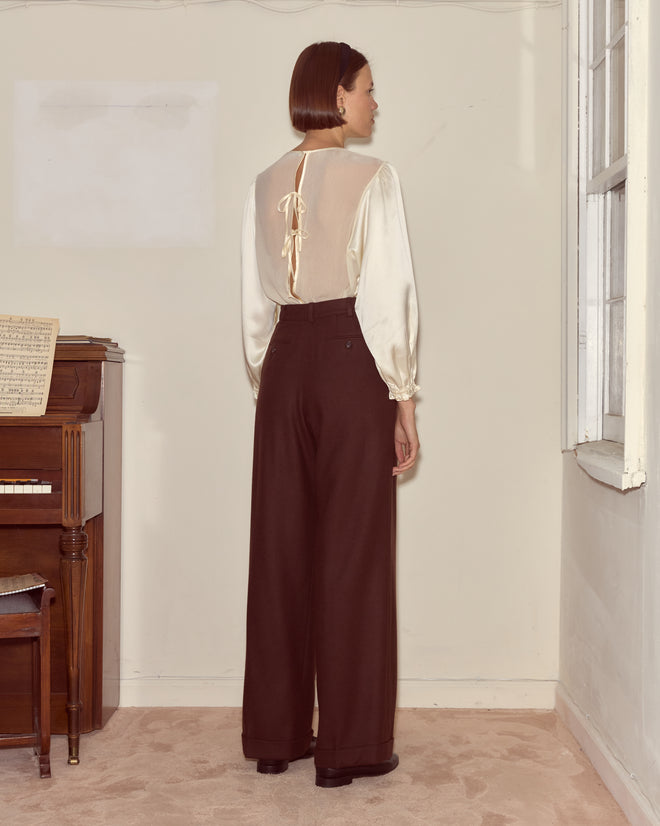 A woman stands indoors near a piano, facing away, wearing a sheer cream blouse paired with Bramble Port Trousers—high-waisted, wide-leg brown wool twill pants.