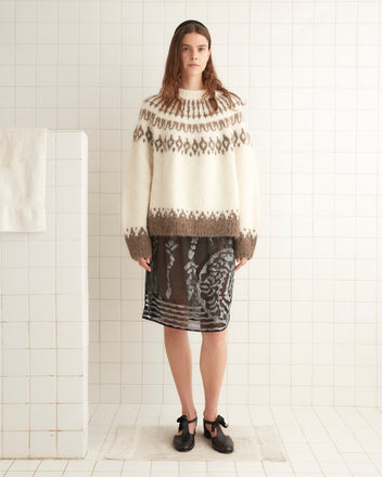 A person stands in a white tiled room wearing a Branch Yoke Sweater, a sheer skirt over a patterned dress, and black shoes.