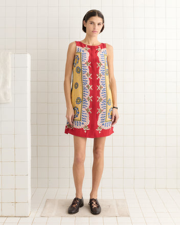 A person wearing a Bricolage Dress, inspired by 1960s patterns in red, stands on a mat in a tiled room with black shoes.