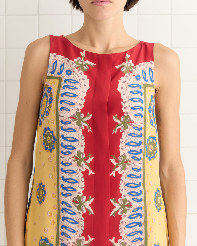 A person stands gracefully in a tiled setting, wearing the Bricolage Dress, a sleeveless piece inspired by the 1960s with vibrant red, yellow, blue, and pink patterned designs.