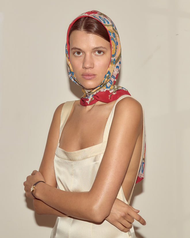 Woman in a cream dress and colorful headscarf stands with arms crossed against a plain background, her look subtly elevated by the Bricolage Driving Cap.