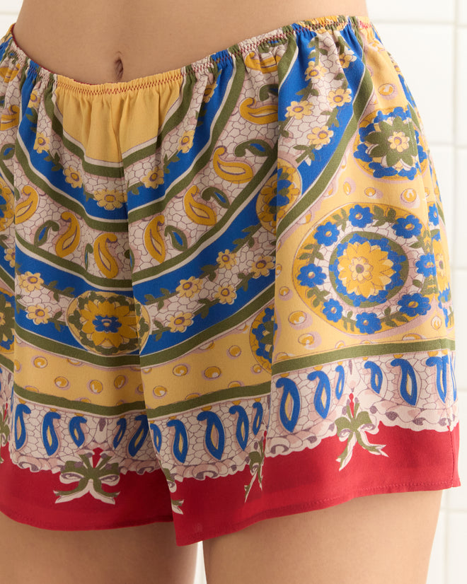 Close-up of a person wearing vibrant Bricolage Shorts, featuring floral and geometric patterns in yellow, blue, and red, reminiscent of 1960s style.