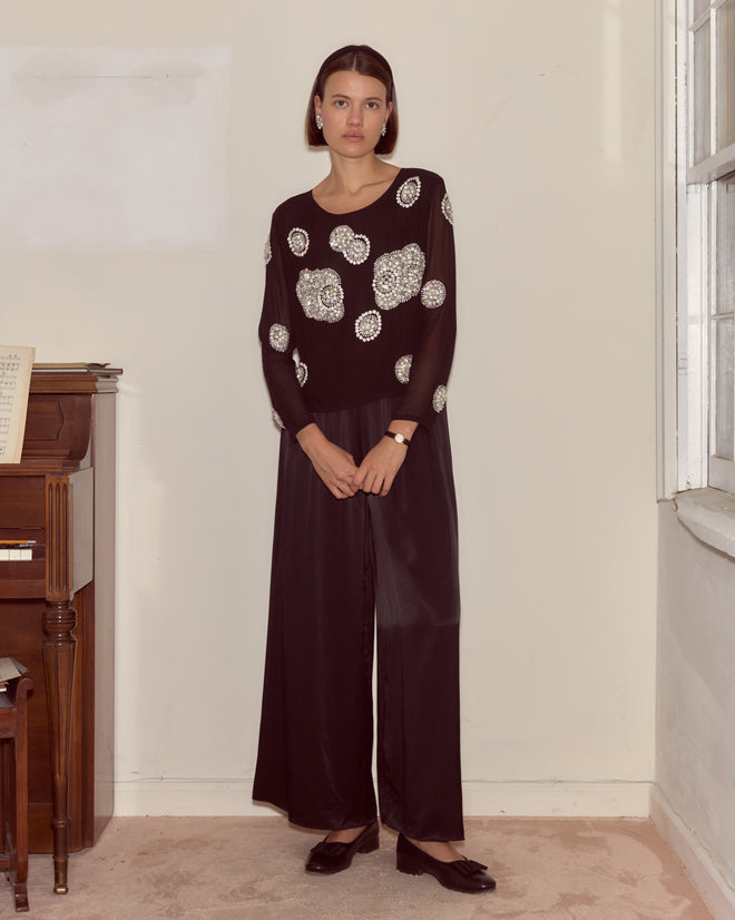 A woman stands indoors by a piano and window, hands clasped, wearing the Brooch Blouse - Black, a silk top detailed with silver sequin circles.