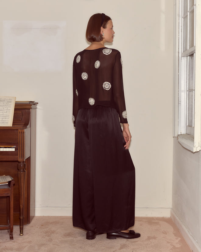 Woman in the Brooch Blouse - Black, featuring sheer fabric with silver circular patterns and delicate hand beadwork, stands near a piano facing away from the camera.