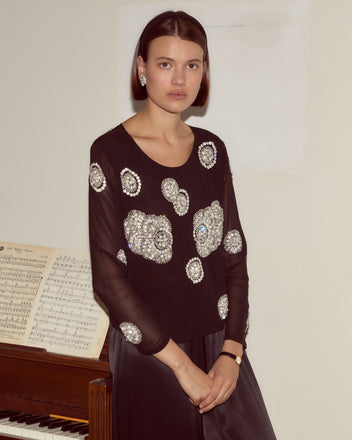 A woman in the black Brooch Blouse stands by a piano with sheet music.